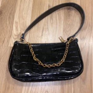 Svart axelväska med kedjedetalj - Snygg svart axelväska med krokodilmönster och guldfärgad kedja. Väskan har en dragkedja upptill och en stilren design som passar perfekt för en kväll ute. Den är kompakt men rymmer det viktigaste.
