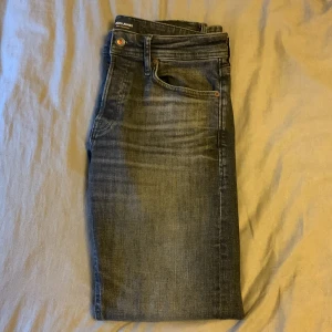 Grå jeans från Jack & Jones - Snygga grå jeans från Jack & Jones med en klassisk femficksdesign. De har en knappgylf och en lätt tvättad look som ger en avslappnad stil. Perfekta för en casual outfit. Jeansen är aldrig använda så skicket är som nytt. Storleken är W30-L32
