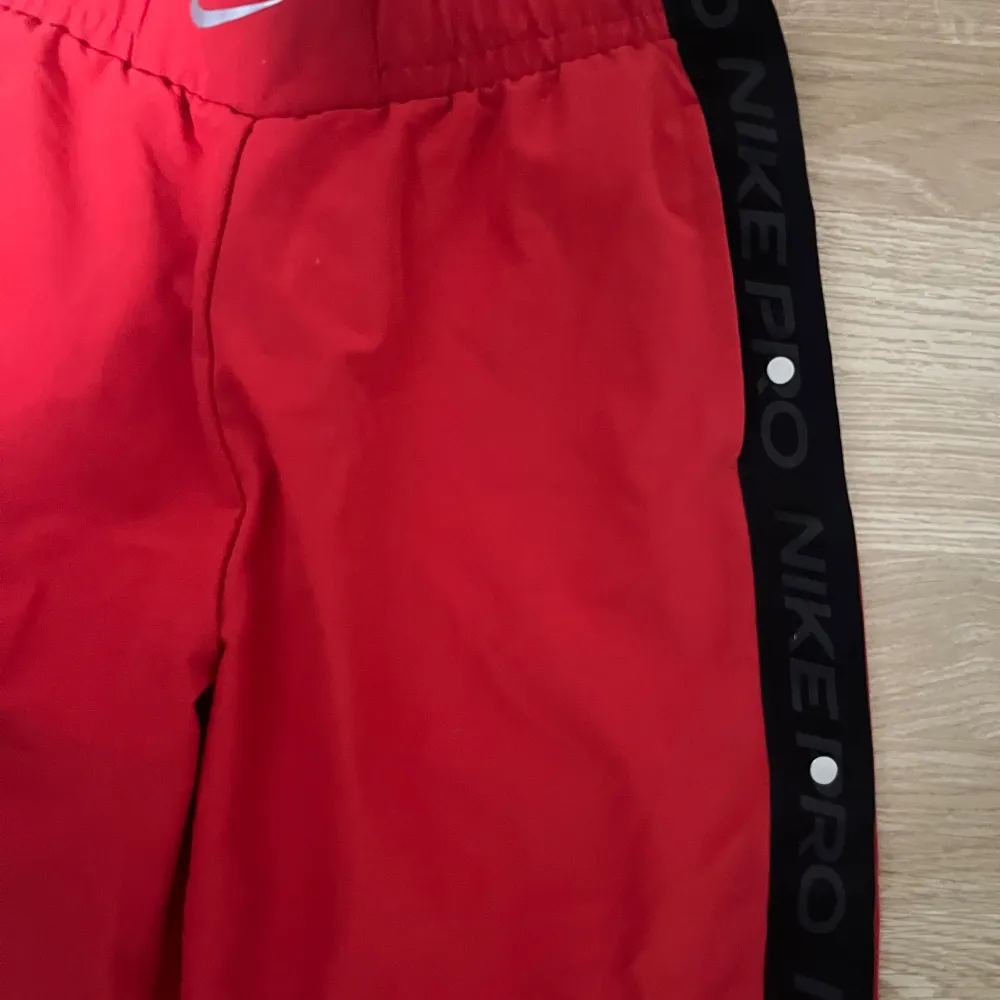 Snygga röda träningsbyxor från Nike med svarta sidopaneler och texten 'NIKE PRO'. Byxorna har en elastisk midja och är perfekta för träning. De har en avslappnad passform och är gjorda i ett lätt material.. Farkut & Housut.