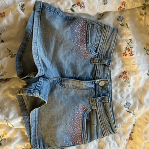 Jeansshorts med rosa pärlor - Snygga jeansshorts med hög midja och dekorativa rosa pärlor vid fickorna. Perfekta för sommardagar och ger en cool touch till din outfit. De har en klassisk jeansblå färg och en bekväm passform. lowwaist