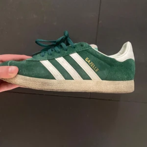 Gröna Adidas Gazelle sneakers - Snygga gröna Adidas Gazelle sneakers i mocka med vita detaljer och snörning. Perfekta för en stilren och sportig look. De ikoniska tre ränderna och Gazelle-loggan i guld ger en klassisk touch. Man får även med box och ett par vita sko snören så man kan ha det man gillar mest