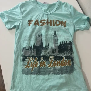 Mintgrön t-shirt med London-tryck - Säljer en mintgrön t-shirt med ett coolt tryck av Londons skyline och texten 'Fashion Life in London'. T-shirten har en v-ringad hals och korta ärmar. Perfekt för dig som älskar London och vill ha en stilren look.