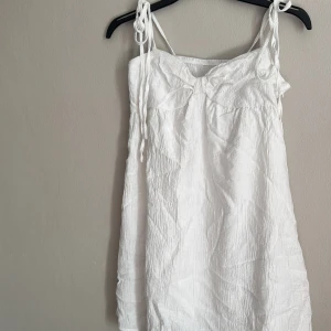 Vit ärmlös klänning - Säljer en söt vit klänning med knytband på axlarna. Klänningen är ärmlös och har en enkel design som passar perfekt för varma dagar. Perfekt för en avslappnad sommarlook!