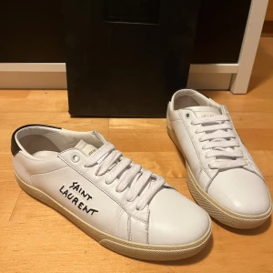 Saint Laurent Court - Snygga vita sneakers från Saint Laurent med svart häl och saint laurent logga på sidan. Skorna har en simpel design med fräscha vita snören och en beige sula. Skorna är 9/10 skick och rekommenderar verkligen. Skorna är i storlek 37 men sitter en storlek större. Passar 37-38,5 Nypris är omkring 7000-7600kr. Tveka inte på att fråga! 