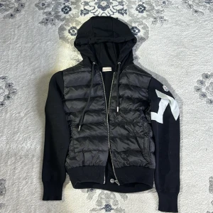 Svart dunväst med huva från Moncler - Snygg svart dunväst från Moncler med huva och dragkedja. Västen har ett coolt mönster på ärmen och ribbade muddar. Perfekt för en stilren look.