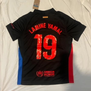 Svart fotbollströja med tryck - Snygg svart fotbollströja från Nike med röda och blå detaljer. Tröjan har korta ärmar och ett stort rött tryck på framsidan. Perfekt för fotbollsfans som vill ha en stilren look.