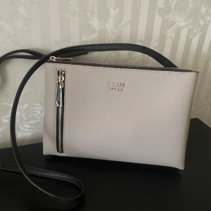 Beige axelväska från Guess - Snygg beige axelväska från Guess med dragkedja och svart axelrem. Knappt använd.