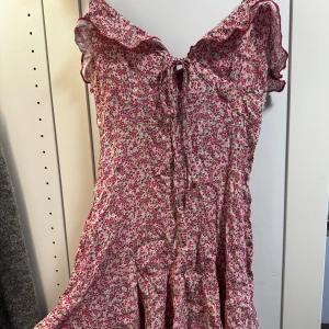 Blommig klänning med volangdetaljer - Charmig blommig klänning i rosa och rött med volangdetaljer och knytning fram. Perfekt för sommarens varma dagar. Klänningen har en dragkedja i ryggen för enkel påklädning. Storlek XS från zara, helt oanvänd!