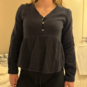 Svart långärmad blus med knappar - Säljer en svart långärmad blus med v-ringning och knappar framtill. Blusen har en lös passform och är tillverkad i ett mjukt material med struktur. Perfekt för en avslappnad stil.