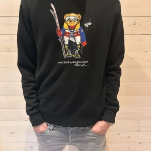 Svart sweatshirt med Polo Bear från Ralph Lauren - Säljer en svart sweatshirt från Ralph Lauren med det ikoniska Polo Bear-motivet på framsidan. Tröjan har långa ärmar och en normal passform. Perfekt för en avslappnad stil med en touch av lyx.
