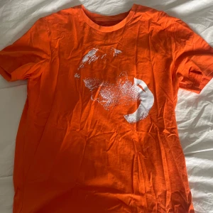 Orange Alec Benjamin t-shirt - Orange t-shirt med vit tryck framtill av Alec Benjamins ansikte och låtlista från albumet (Un)Commentary på ryggen. Klassisk passform med rund hals och korta ärmar. Perfekt för dig som gillar musikmerch och vill sticka ut med färg.