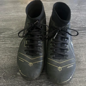 Svarta Nike fotbollsskor med guldiga detaljer - Snygga svarta fotbollsskor från Nike med guldiga linjer och detaljer. Skorna har snörning, högre skaft och mönstrad ovansida för extra grepp. Sulan har fasta dobbar för bra fäste på planen. Perfekta för dig som vill sticka ut på matchen.