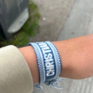 Blått Christian Dior J'Adior armband - Säljer ett snyggt blått tygarmband från Christian Dior med texten 'J'ADIOR'. Armbandet har vita detaljer, knytning med tofsar och är perfekt för dig som vill ha en trendig accessoar med lyxig känsla.