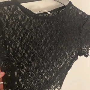 Svart spetsig croppad topp FB Sister - Svart croppad topp från FB Sister i storlek XS. Hela toppen är i genombruten spets med korta ärmar och rund halsringning. Perfekt för att ge outfiten en edgy och trendig vibe.