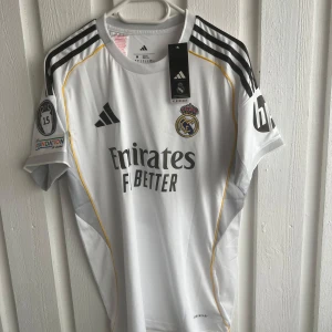 Real Madrid Mbappé 9 matchtröja Adidas - Snygg vit Real Madrid fotbollströja från Adidas med Mbappé och nummer 9 på ryggen. Tröjan har svarta detaljer, gula linjer och klubbmärke på bröstet. Korta ärmar, officiella tryck och Champions League-märke på ärmen. Tillverkad i lätt funktionsmaterial.