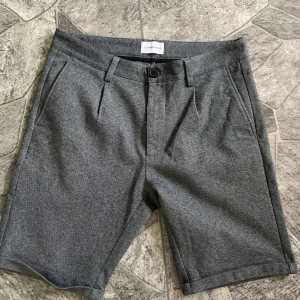 Grå shorts från Lindbergh White M - Stilrena grå shorts från Lindbergh White i klassisk modell med knapp och dragkedja framtill, bälteshällor och fickor både fram och bak. Perfekta för en clean look under varmare dagar. Materialet känns mjukt och har en lätt struktur.