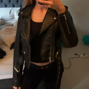 Svart skinnjacka med dragkedjor - Snygg svart skinnjacka med klassisk bikerstil, silverfärgade dragkedjor och bälte i midjan. Jackan har långa ärmar och coola detaljer som ger en edgy vibe. Perfekt för dig som vill ha en tidlös och trendig look.