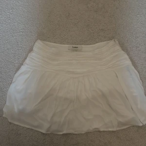 Vit kortkjol från Pull&Bear - Superfin vit kortkjol från Pull&Bear med broderade detaljer och lätt volang. Kjolen har en luftig känsla och är perfekt för varma dagar. Snyggt rynkad upptill och i mjukt material som känns skönt mot huden.