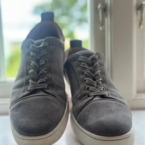 Grå mockasneakers från Christian Louboutin - Snygga grå sneakers i mocka från Christian Louboutin med klassisk vit sula och ikonisk röd undersida. Skorna har rund tå, snörning och diskreta metalldetaljer vid snörhålen. Perfekta för dig som vill ha en lyxig och stilren look. Just nu är dom lite smutsiga men ska lämna in på skotvätt innan dom skickas. Kom privat för flera bilder