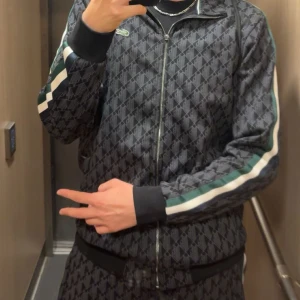 Svart mönstrad track jacket Lacoste och byxor - Svart track jacket från Lacoste med diskret geometriskt mönster över hela jackan. Har dragkedja framtill, ståkrage och vita samt gröna ränder längs ärmarna. Klassisk krokodillogga på bröstet. Perfekt för en sportig och stilren look.
