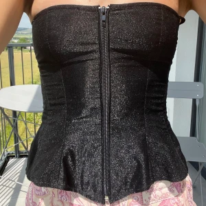 Glittrig svart korsett med snörning - Säljer en svart glittrig korsett med dragkedja framtill och snygg snörning i ryggen. Toppen är axelbandslös och har en lätt utsvängd nederkant som framhäver midjan ännu mer. Perfekt för dig som älskar snygga och bekväma outfits.