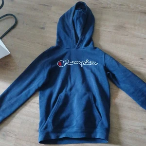 Mörkblå Champion hoodie  - Säljer en mörkblå hoodie från Champion med stor broderad logga på bröstet. Tröjan har huva, känguruficka och långa ärmar. Perfekt för dig som gillar sportig och avslappnad stil. Pris kan diskuteras 