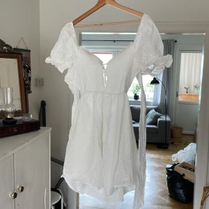 Klänning - Superfin vit kort klänning med puffärmar och v-ringning. Klänningen har broderade spetsdetaljer, smockad överdel och knytning framtill. Perfekt för sommarens alla tillfällen och ger en romantisk vibe.