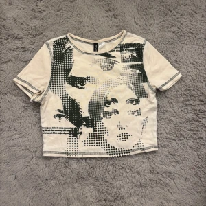Beige croppad topp med ansiktsprint - Cool croppad topp från Divided i beige med svart grafiskt ansiktsmönster framtill. T-shirten har korta ärmar, kontrastsömmar och en tight passform. Perfekt för dig som gillar edgy och unik stil.