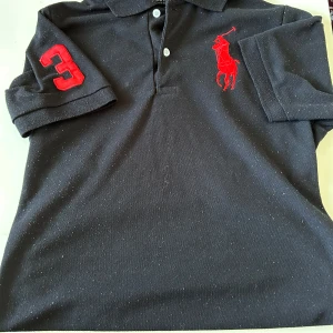 Svart pikétröja från Ralph Lauren - Svart pikétröja från Ralph Lauren med röd broderad logga på bröstet och en stor röd siffra 3 på ärmen. Klassisk krage, korta ärmar och knäppning framtill. Tillverkad i bomull och har en sportig vibe.