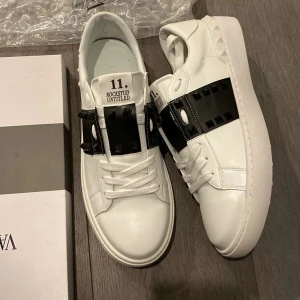 Valentino Rockstud vita sneakers - Snygga vita Valentino Rockstud sneakers med svart band och ikoniska nitar över vristen. Skorna har rund tå, platt sula och är gjorda i skinn. Perfekta för dig som vill sticka ut med en lyxig och edgy stil.