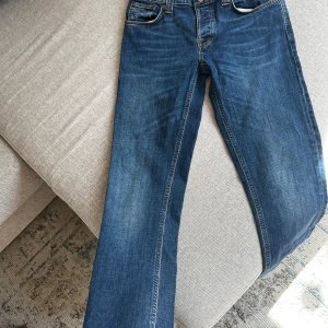 Blå jeans från Nudie Jeans Co 31/34 - Säljer ett par klassiska lågmidja  blå jeans från Nudie Jeans Co i storlek 31/34. Jeansen har raka ben, knappgylf och snygga detaljer på bakfickorna. Tillverkade i 100% ekologisk bomull med orangea sömmar och en cool tvättad look. Nyskick. 