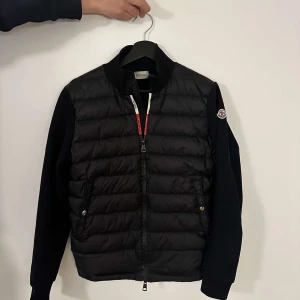 Svart pufferjacka från Moncler - Snygg svart pufferjacka från Moncler med quiltad framsida och släta ärmar. Jackan har dragkedja framtill, ribbad krage och två fickor med knapp. Klassisk Moncler-logga på ärmen och coola detaljer vid dragkedjan. Perfekt för dig som gillar stilren och sportig look.