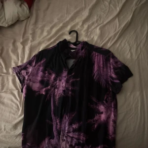 Lila mönstrad skjorta Avenue Tropicale - Säljer en kortärmad skjorta från Avenue Tropicale i storlek medium. Skjortan har ett tropiskt mönster i lila och svart med palmer, knäppning framtill och avslappnad passform. Perfekt för dig som vill sticka ut med en cool vibe.