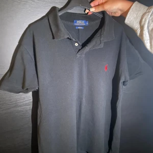 Svart pikétröja från Polo Ralph Lauren - Klassisk svart pikétröja från Polo Ralph Lauren i bomull. Tröjan har korta ärmar, krage med knappar och den ikoniska röda broderade loggan på bröstet. Custom fit-modell som sitter snyggt och passar till det mesta. Priset är inte hugget i sten och kan enkelt diskusar vid smidig affär!