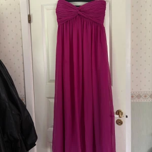 Axelbandslös cerise långklänning - Snygg axelbandslös långklänning i cerise med twistad överdel och vackert fall. Perfekt för bal eller fest, med en luftig och elegant känsla. Klänningen har en romantisk look och är lätt att matcha med smycken och klackar.