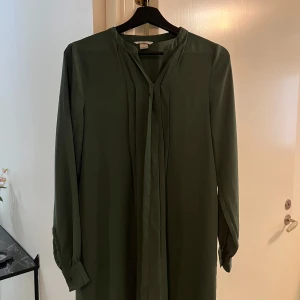 Mörkgrön långärmad klänning - Stilren mörkgrön klänning med lång ärm och knappar vid ärmslut. Rak passform och enkel design med ett smalt knytband vid halsen. 