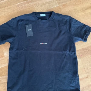 Svart Saint Laurent t-shirt i bomull - Svart t-shirt från Saint Laurent med minimalistisk logga broderad på bröstet. Klassisk rund halsringning och korta ärmar. Tillverkad i mjuk bomull som känns skön mot huden. Perfekt för dig som gillar stilrena designer och exklusiva märken. Självklart äkta✨👌
