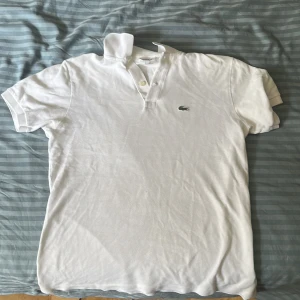 Vit pikétröja från Lacoste - Klassisk vit pikétröja från Lacoste med korta ärmar och den ikoniska gröna krokodilloggan på bröstet. Tröjan har krage, knappar framtill och är tillverkad i bomull som känns luftig och skön. Perfekt för en clean och sportig stil.