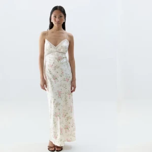 Vit blommig långklänning i satin med spets - Helt ny romantisk långklänning i vit satin med tunna axelband och v-ringning. Klänningen har ett sött blommigt mönster i rosa och grönt samt spetsdetaljer vid bysten. Perfekt för sommarens alla tillfällen.