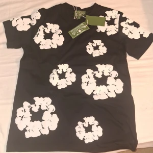 Svart blommig t-shirt från Denim Tears - Svart t-shirt från Denim Tears med stora vita blommiga tryck över hela plagget. Klassisk passform med rund hals och korta ärmar. Materialet är mjuk bomull som känns skönt mot huden. Perfekt statement-piece för dig som vill sticka ut.