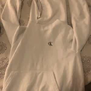Vit hoodie från Calvin Klein - Säljer en stilren vit hoodie från Calvin Klein med broderad CK-logga på bröstet. Hoodien har huva med snörning, magficka och ribbade muddar vid ärmslut och midja. Perfekt för dig som gillar en clean och enkel look.