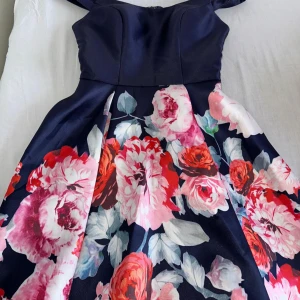 Mörkblå blommig kortklänning med axelband - Supersöt mörkblå kortklänning med breda axelband och voluminös kjol. Klänningen har stora blommor i rosa, rött och vitt med gröna och blå blad. Materialet är glansigt och känns lyxigt, perfekt för att sticka ut. Kjolen har fina veck och toppen är figurnära.