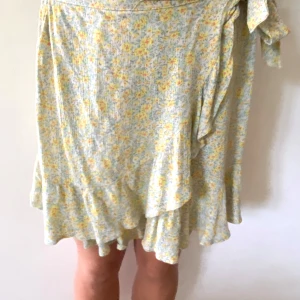 Blommig omlottkjol med volang - Supersöt kort omlottkjol med volangkant och blommigt mönster i ljusgult och vitt. Kjolen har knyt i midjan och är perfekt för varma dagar. 