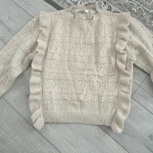 Beige stickad tröja med volang från by clara - Supersöt beige stickad tröja från by clara med volangdetaljer längs axlarna och ett snyggt hålmönster. Tröjan har lång ärm och ribbade muddar vid hals, ärmslut och nederkant. Perfekt för dig som gillar romantisk och trendig stil. Köpt från missöy Gbg. Nyskick