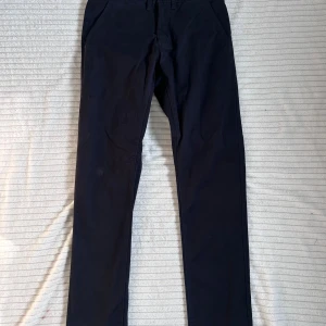Svarta chinos från Dressmann slim fit - Svarta chinos från Dressmann i slim fit-modell. Klassisk design med snedställda fickor fram och två bakfickor med knapp. Byxorna har bälteshällor och stängs med knapp och dragkedja. Perfekta för en clean och stilren look.