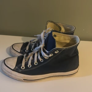 Mörkblå Converse All Star high-tops - Klassiska mörkblå Converse All Star high-tops med vit sula och vita snören. Skorna har rund tå, tydliga kontrastsömmar och den ikoniska loggan på sidan. Canvasmaterial och platt sula gör dem till en tidlös favorit för alla stilar.