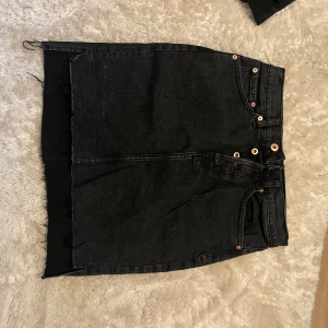 Svart jeanskjol från 157, stl S - Cool svart jeanskjol från 157 med råa kanter och klassiska fickor både fram och bak. Kjolen har knapp- och dragkedjestängning samt detaljer i kopparfärg. Perfekt för dig som gillar en edgy och avslappnad stil.