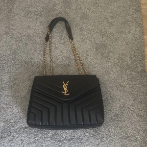 Svart YSL axelväska i skinn - Säljer en svart quiltad axelväska från YSL med ikoniskt guldigt YSL-emblem framtill. Väskan har kedjeremmar i guld och är tillverkad i mjukt skinn. Snygg och stilren design med klassiska detaljer och magnetlås. Perfekt för dig som vill ha en lyxig väska med statement.