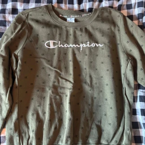 Olivgrön Champion sweatshirt XL - Olivgrön sweatshirt från Champion i storlek XL. Tröjan har ett broderat Champion-logo på bröstet och små svarta Champion-loggor över hela tyget. Mjuk bomullskänsla, rund hals och långa ärmar. Perfekt för en chill och sportig stil.