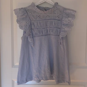 Ljusblå blus med spets från Lindex - Supersöt ljusblå blus från Holly & Whyte by Lindex. Blusen har broderade spetsdetaljer upptill och på de volangiga ärmlösa axlarna, samt rynkade partier framtill. Perfekt för en fräsch och somrig look!